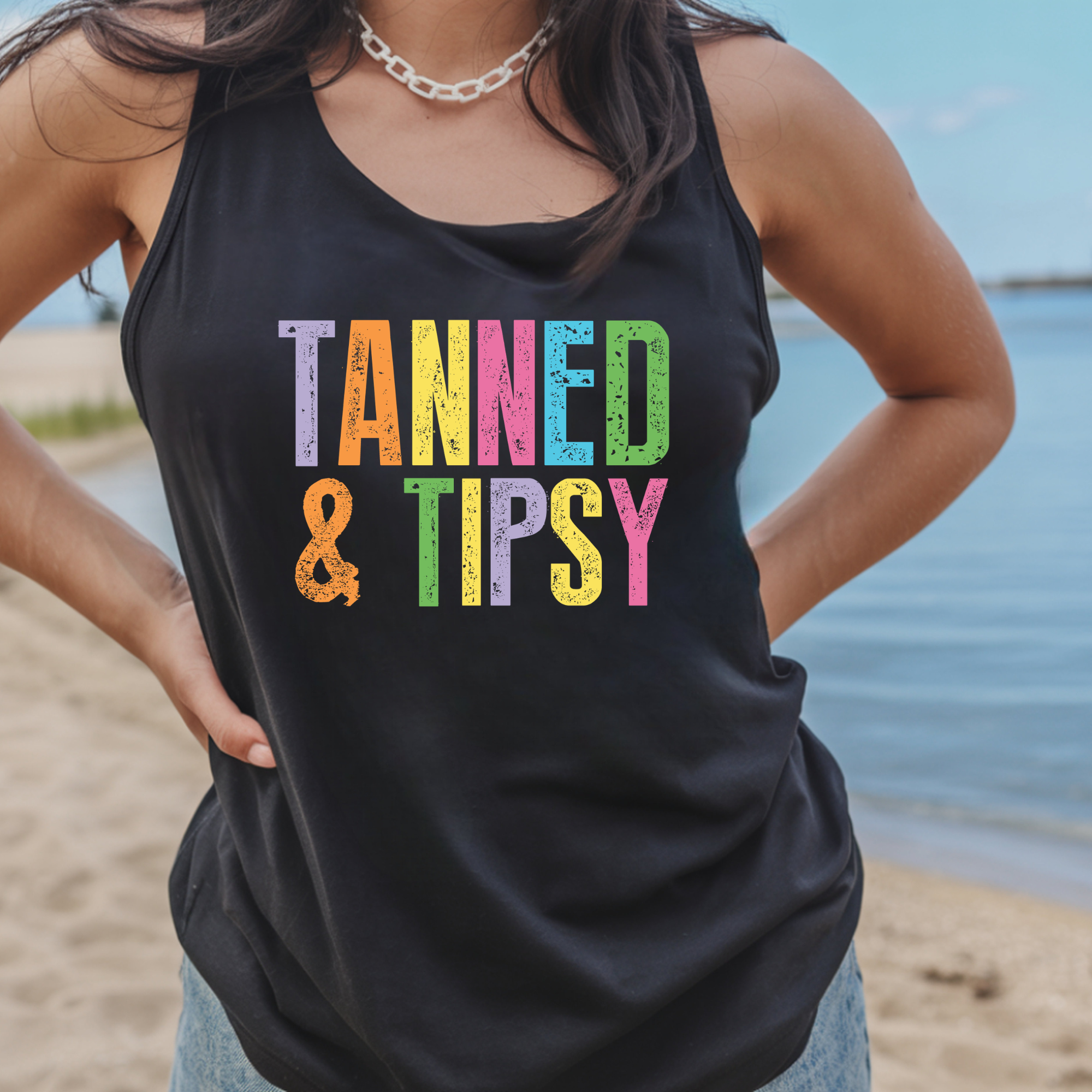 Person wearing a black tank top with 'Tanned & Tipsy' text at the beach